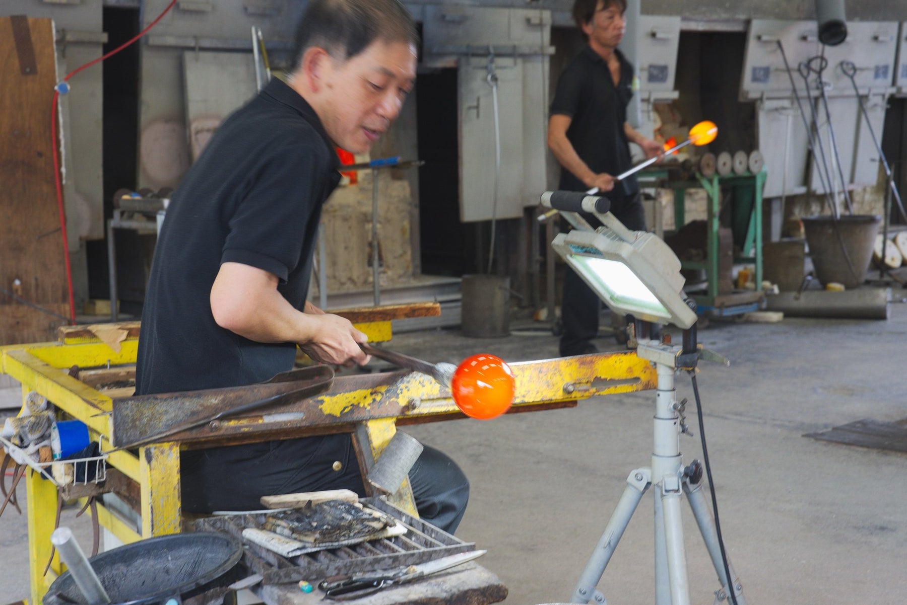 Two glassblowers shaping molten glass at furnace stations