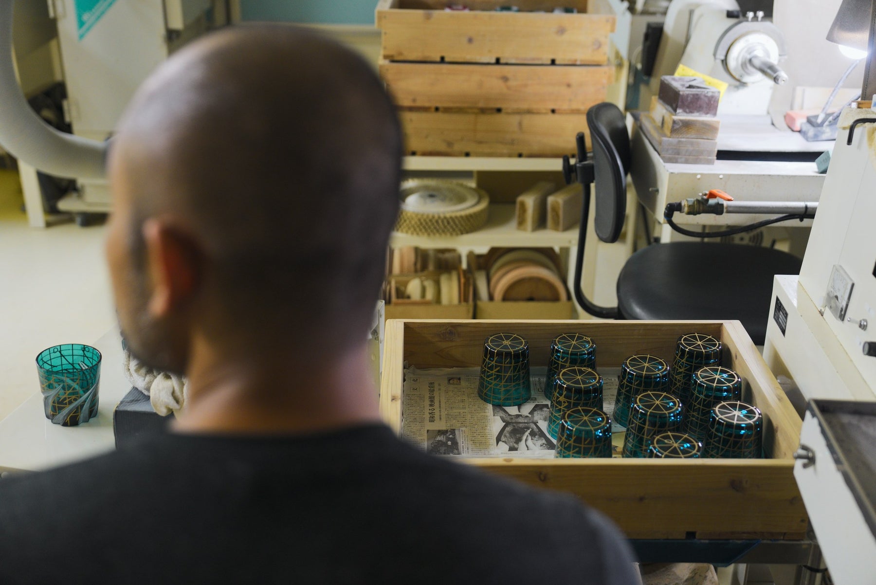 Craftsman at a cutting-wheel bench beside a crate of teal tumblers
